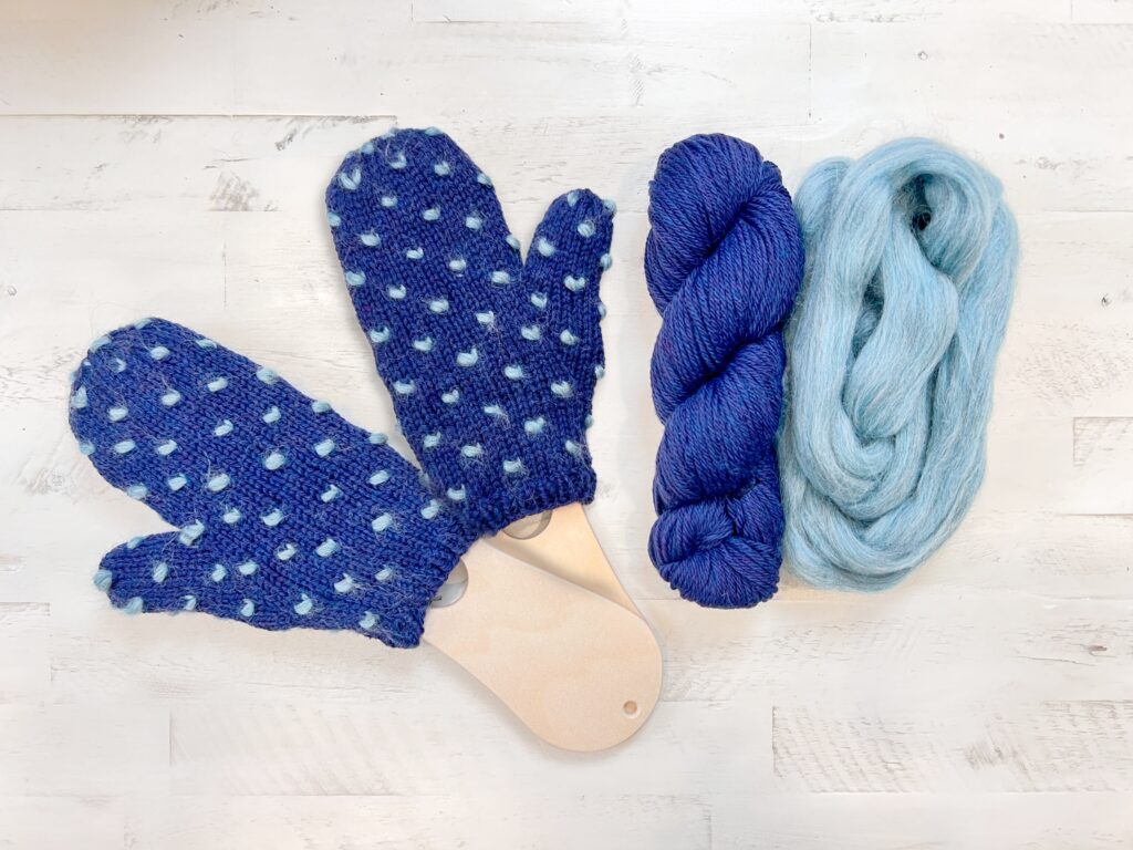 Two blue knitted Andes Thrummed Knit Mittens with light blue dots are displayed on wooden mitten blockers. Beside them are a skein of dark blue yarn and a bundle of light blue roving, perfect for the First Fall 2025 Make-Along Free Knitting Pattern. -Marly Bird