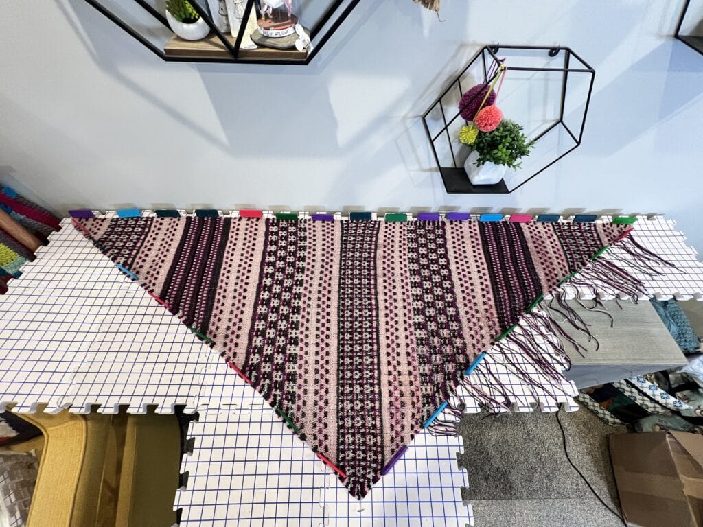 Striped mosaic corner to corner triangle knit shawl in purple, black, and cream, blocked on a gridded matt stitch texture clearly visible.