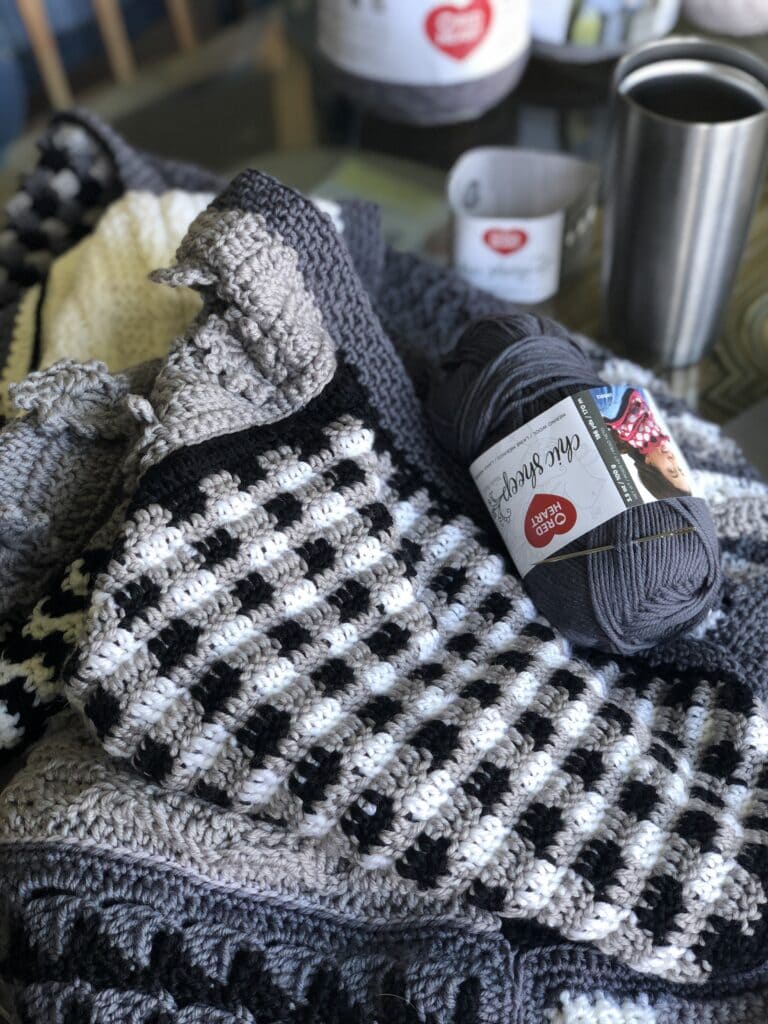 A pile of handmade crocheted blankets in shades of gray, black, and white, with a skein of gray yarn labeled "Red Heart Chic Sheep" resting on top. A stainless steel travel cup and a coffee mug sit nearby, perfect for cozying up with your favorite podcast. -Marly Bird