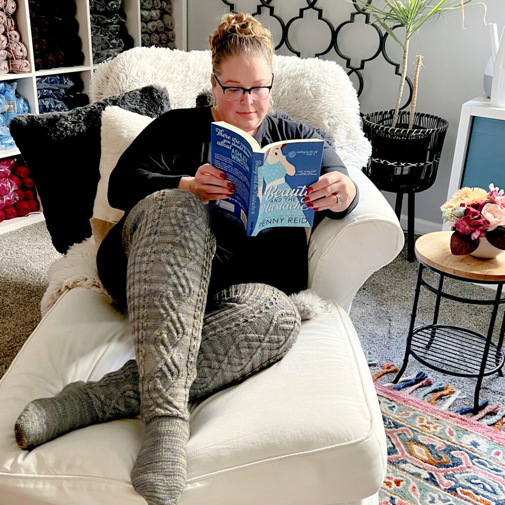 A person with glasses and hair in a bun is sitting on a white chair, reading a book titled "Beauty and the Mustache." They're wearing a black shirt and thigh high knit socks. The cozy room has yarn shelves, a plant, and a colorful rug. -Marly Bird