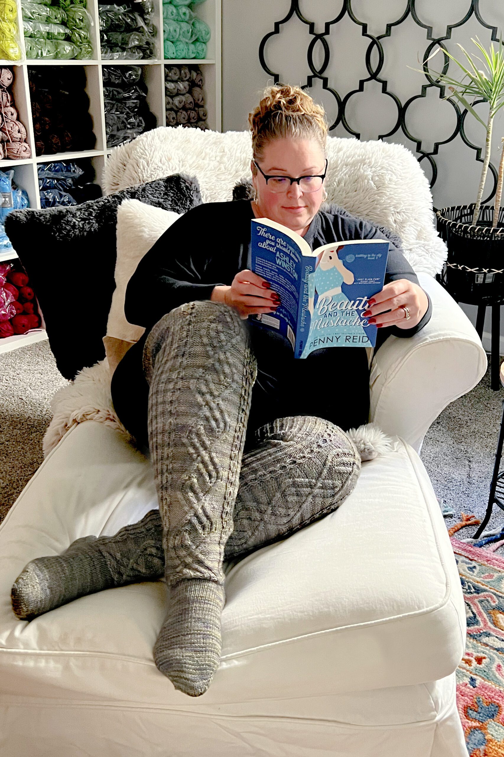 A person with glasses and a bun hairstyle is seated in a white armchair, reading "Beauty From Ashes" by Jenny Reid. Dressed in a black outfit with long knitted socks, they are surrounded by yarn organized on shelves, capturing the cozy essence of Marly Bird's tutorial space amidst modern decor. -Marly Bird
