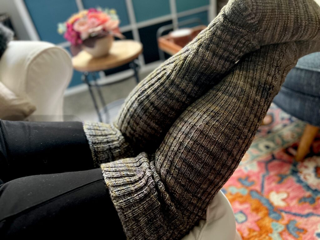 A person in black pants wearing cozy, chunky knit socks with shades of green and gray rests their feet up on a white, cushioned surface. The background includes colorful decor, including a vibrant, patterned rug and a side table with a flower arrangement. It’s the perfect setting to Appalachian Cozy Up. -Marly Bird