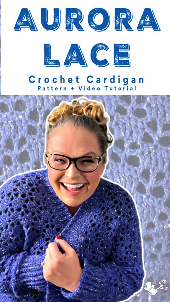 Smiling Marly Bird wearing glasses and a cobalt blue crochet cardigan with intricate lace detailing, embracing herself with one hand while standing against a home background with bookshelves.