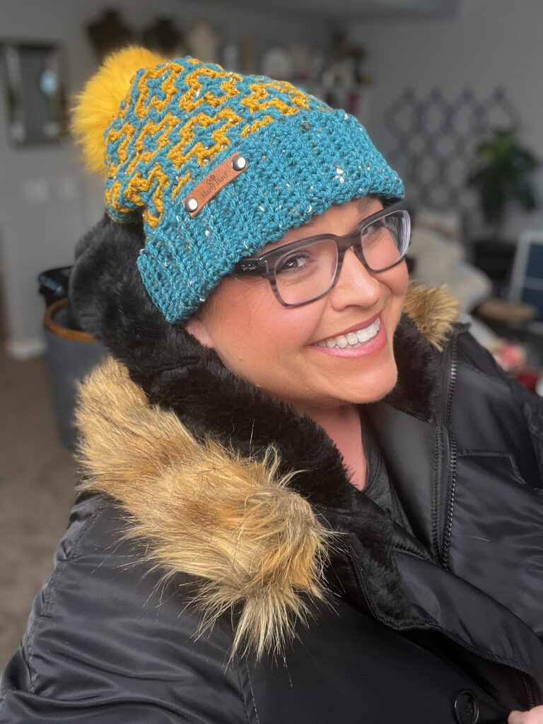 A person smiles warmly, wearing glasses, a Crochet Mosaic Hat in teal with a yellow pattern and pom-pom, and a black coat with a faux fur hood. The background is an inviting indoor space with softly blurred details. -Marly Bird