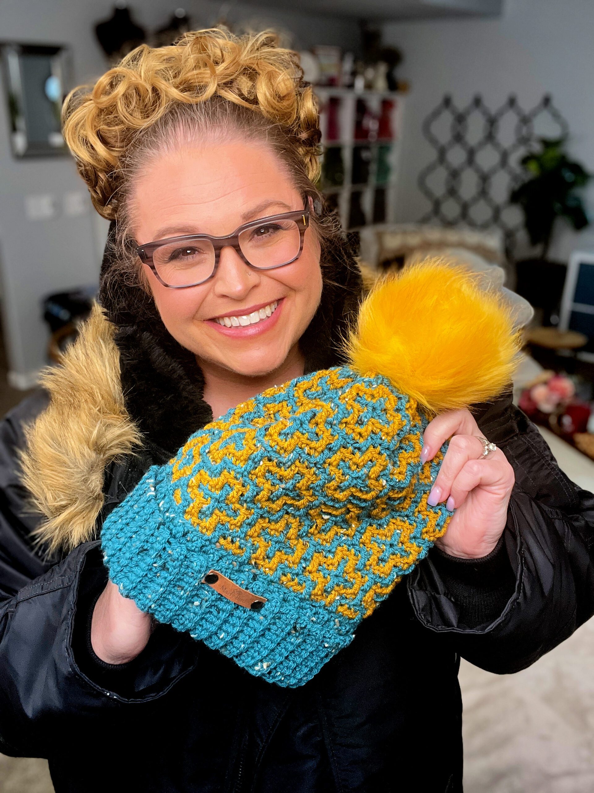 A person with curly hair and glasses smiles while holding an Autumn Breezes Crochet Hat, showcasing its knitted blue and yellow star pattern with a cheerful pom-pom. They are wearing a jacket with a faux fur collar and standing in a cozy room. -Marly Bird