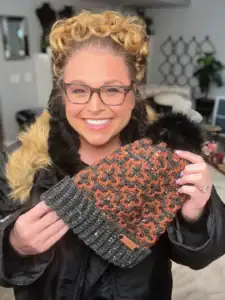 A smiling woman in glasses and a black coat with a fur collar holds up an orange and gray knitted hat with a black pom-pom, standing indoors—perfect inspiration for those seeking free knitting and crochet patterns. -Marly Bird