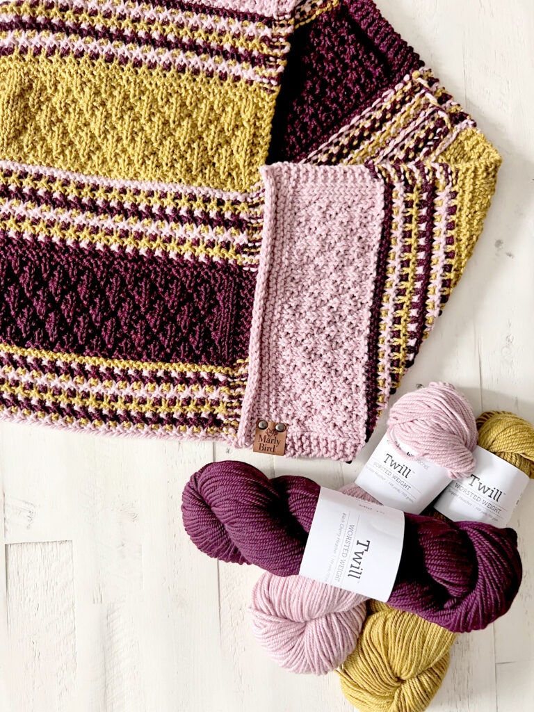 Flat lay image showcasing the Bamberg Bandana Cowl in progress with rich textures in pink, yellow, and purple yarns. The knitting project is laid out on a white surface, displaying its intricate patterns and a Marly Bird branded label. Beside the cowl, there are three skeins of Twill yarn from WeCrochet in coordinating colors, indicating the high-quality materials used in the creation of this piece.