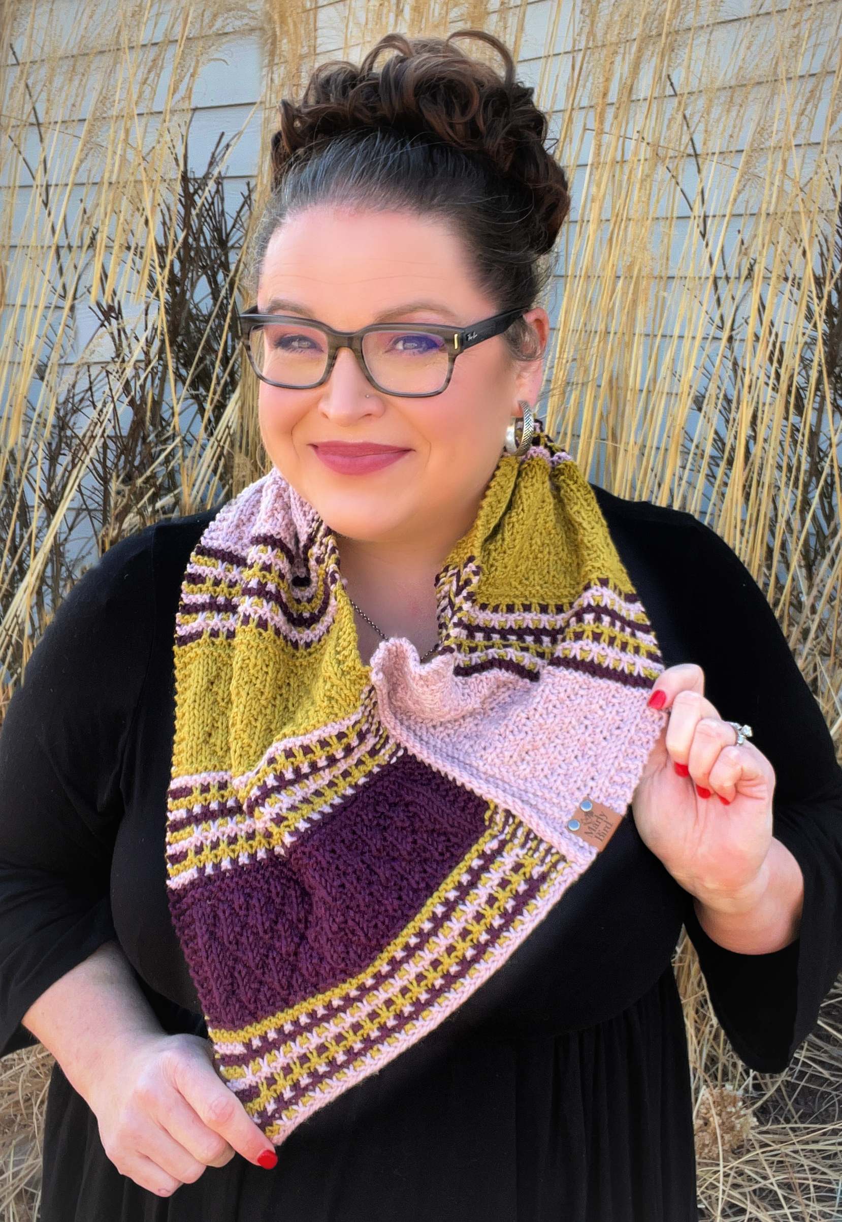 Alt text: "Marly Bird showcasing the Bamberg Bandana Cowl, posing confidently outdoors with tall, golden grass in the background. The cowl features a striking combination of yellow, purple, and pink hues with rich textures. Marly is smiling, wearing glasses, and has her hair styled in an updo. The cowl is artfully draped to display the intricate stitch patterns and a small branded label with 'Marly Bird' is visible on the edge of the cowl.