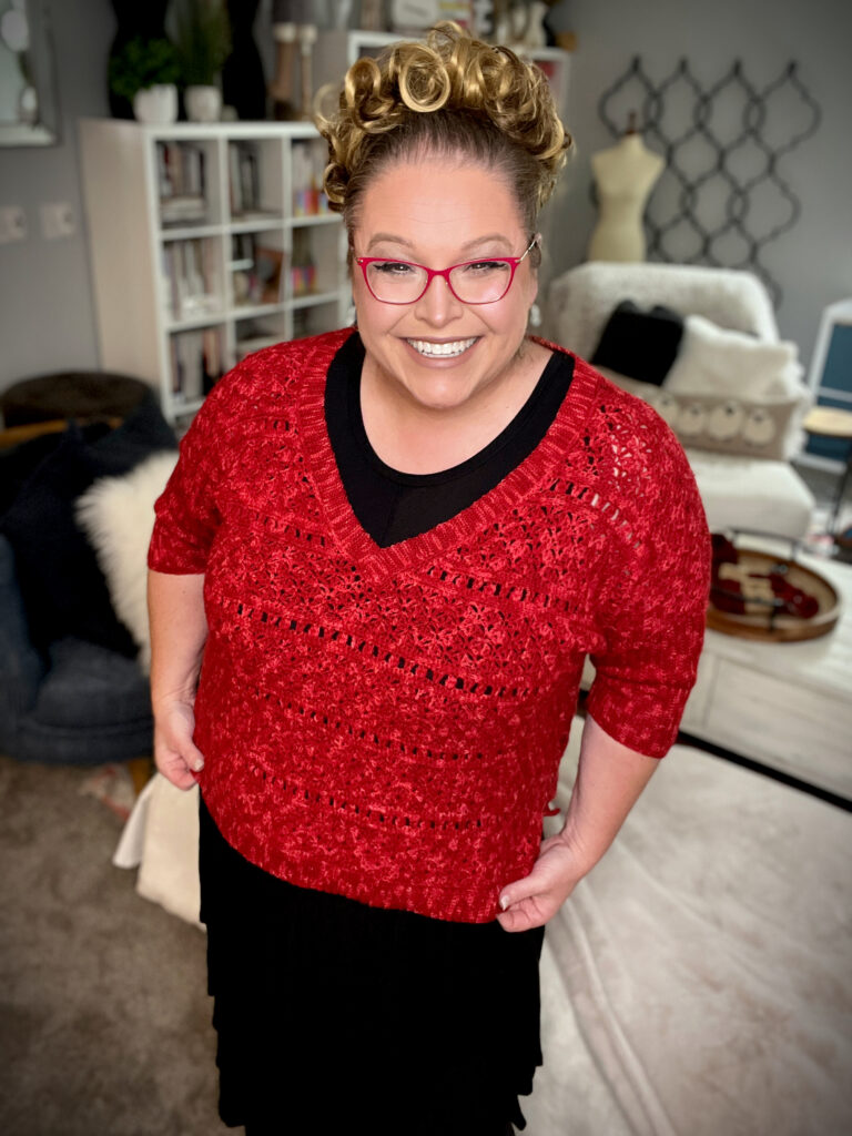 A smiling person with curly blonde hair, wearing red glasses and a bellini crochet sweater over a black dress, stands in a cozy room with a bookshelf and various furniture pieces in the background. -Marly Bird