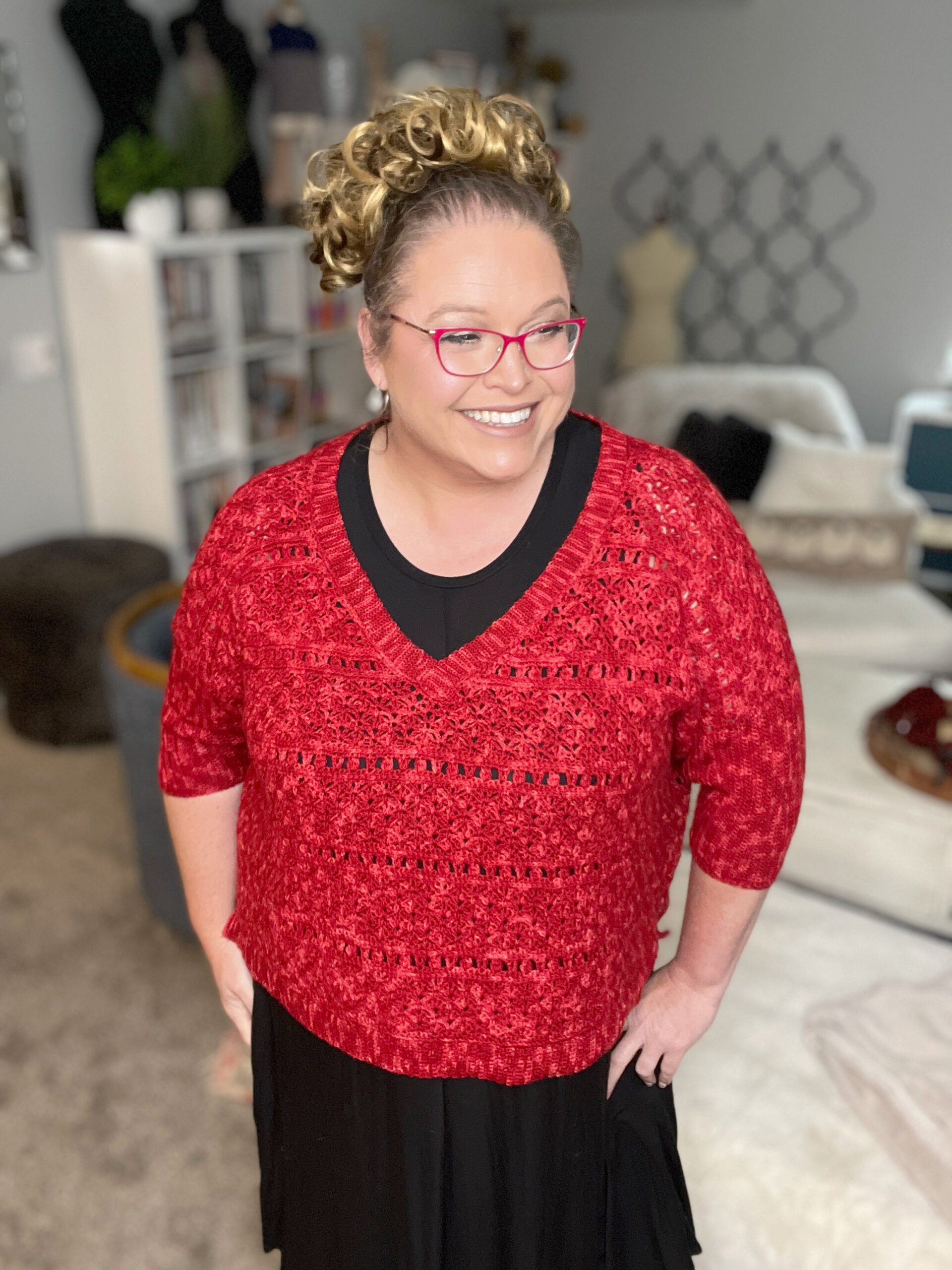 Marly Bird is wearing red glasses, a bellini crochet sweater over a black top, and a black skirt stands in a cozy, well-decorated room with a bed and various home decor items in the background. -Marly Bird