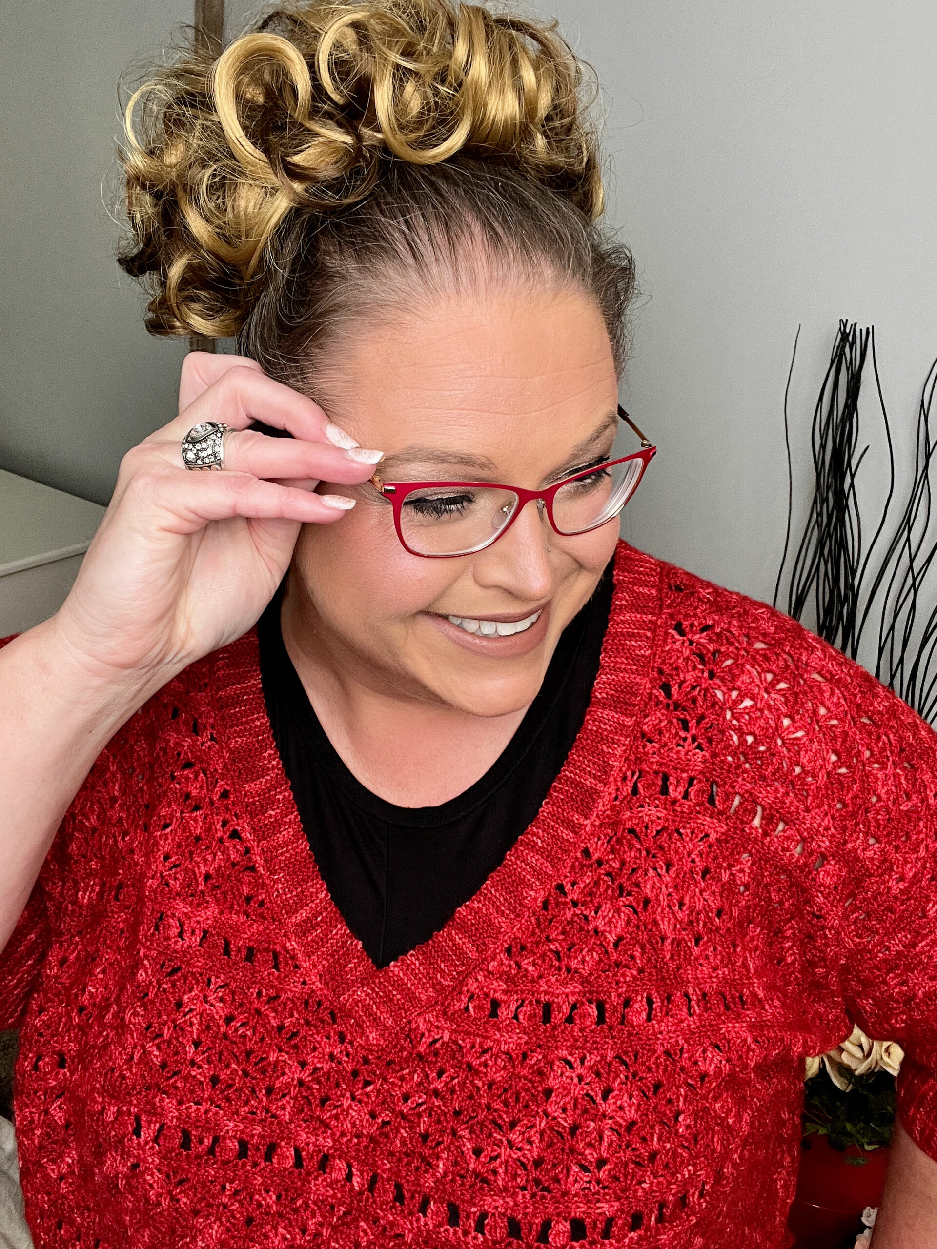 A woman with curly blonde hair styled up is smiling and looking to the side. She is wearing red glasses and a cozy knit sweater over a black top. She has a ring on her right hand, which is touching her glasses. The neutral background features some decorative elements that add charm. -Marly Bird