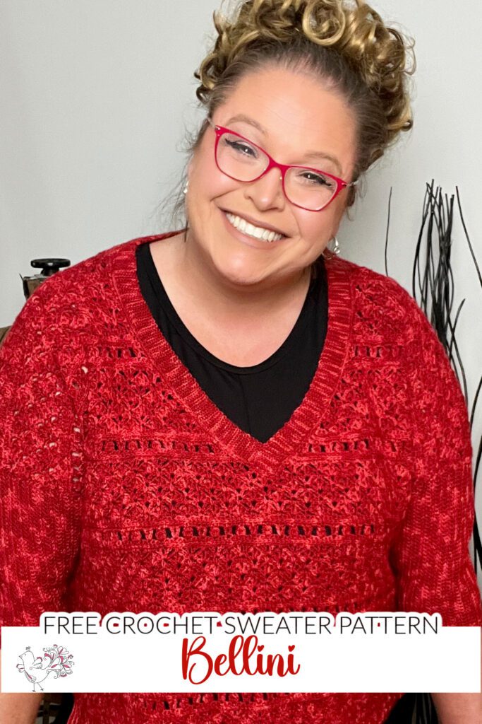 A smiling person with curly hair and red glasses is wearing a bellini crochet sweater. At the bottom of the image, there's text that reads "FREE CROCHET SWEATER PATTERN: Bellini." A black thread stand and some decor are in the background. -Marly Bird