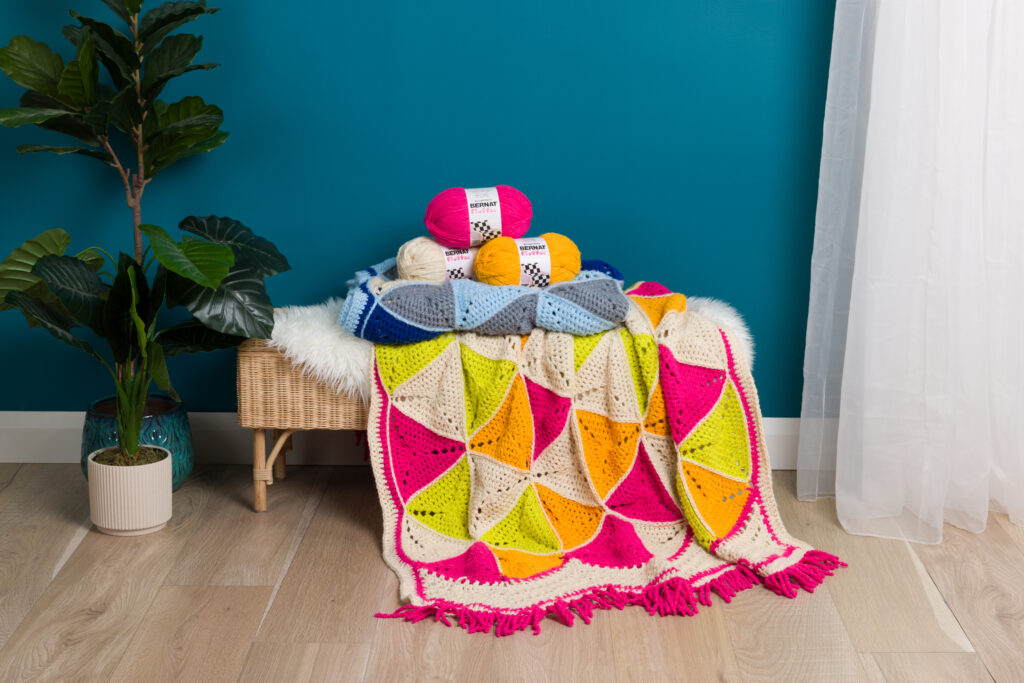 A cozy, colorful crochet blanket with vibrant triangular patterns and fringed edges is draped over a small wicker bench. Balls of yarn in various colors are stacked on the bench. A potted plant stands to the left, and sheer white curtains hang to the right.