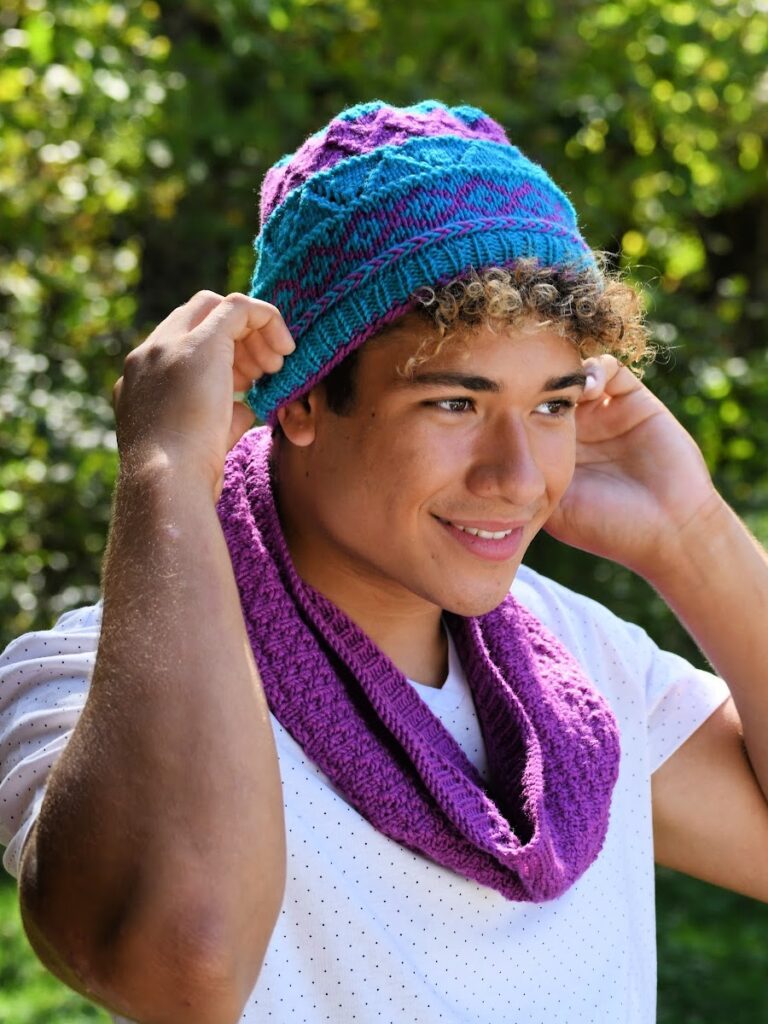 The image features a smiling individual outdoors adjusting a vibrantly colored knitted hat. The hat displays a dynamic colorwork pattern in shades of blue and pink, with a ribbed edge providing a snug fit. The person is also wearing a cowl around their neck in a complementary shade of purple, suggesting the items could be part of a matching set. The natural lighting and greenery in the background accentuate the vivid colors of the knitwear, highlighting the craftsmanship and style of the accessories. The overall image conveys a sense of casual fashion and the joy of wearing personally crafted items. - pattern is BiCrafty Bootcamp My First Knit Hat and Complimentary Cowl by Marly Bird






