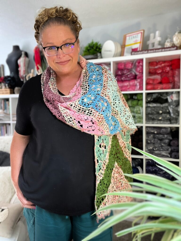 A woman with curly hair and glasses smiles while wearing a colorful, lacy knit shawl. She stands in a room filled with yarn and crafting supplies, perfect for exploring Crochet Shawl Patterns inspired by Marly Bird. A green plant sits in the foreground. -Marly Bird
