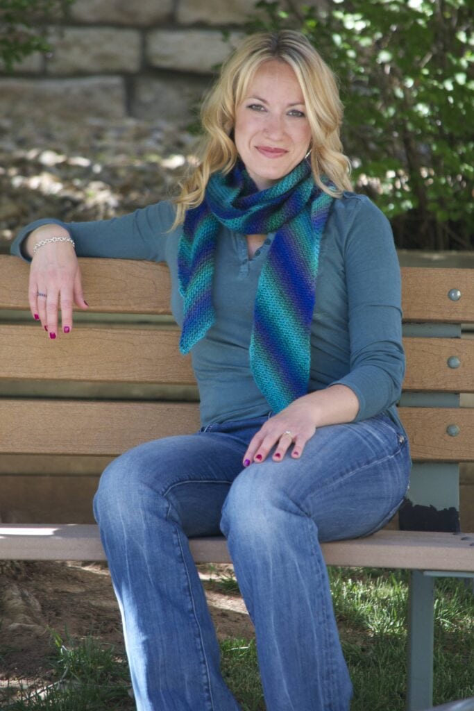 Woman models a blue-green garter stitch shawl, showing its soft texture while seated on a wooden bench outdoors.