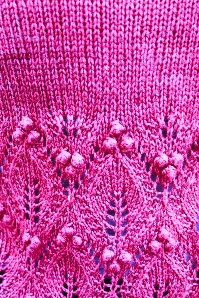 Close-up of pink knitted fabric with a decorative lace pattern and raised bobble stitches, showing intricate texture and detail. -Marly Bird