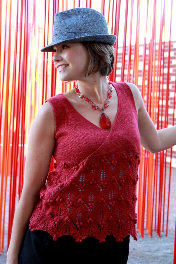 A smiling woman in a gray fedora, red sleeveless knit top with lace details, and a red pendant necklace stands in front of hanging red ribbons, looking to the side. -Marly Bird
