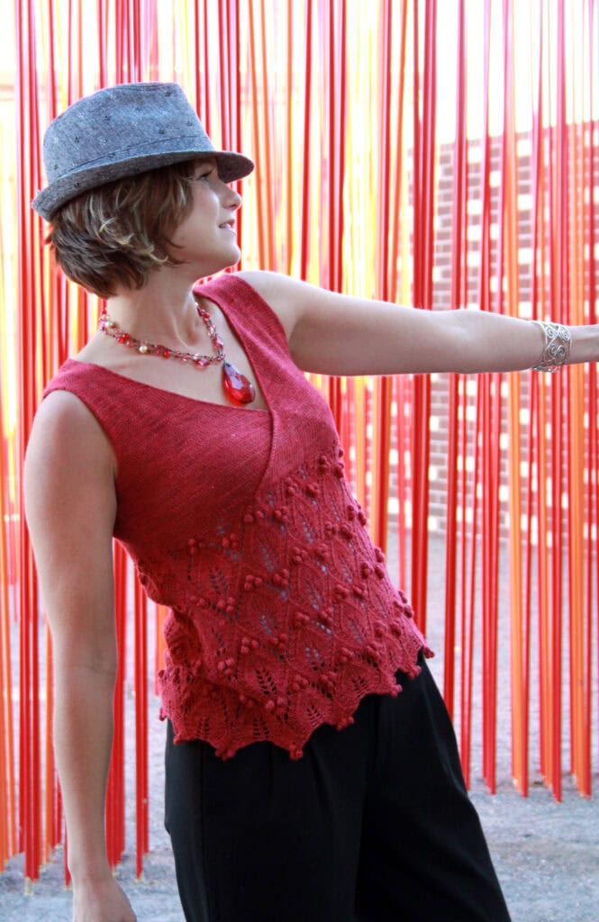 A woman wearing a gray hat, red sleeveless knit top with a lacy pattern, black pants, and red jewelry stands in front of hanging orange ribbons, looking to the side with one arm extended. -Marly Bird