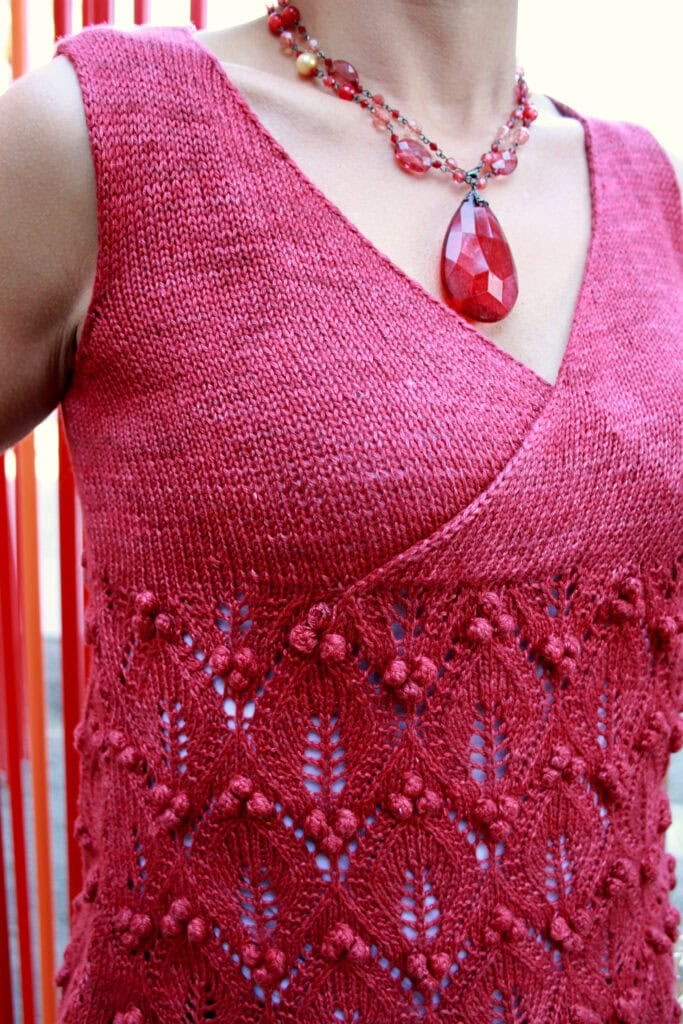 A person wearing a sleeveless, deep pink knit top with a textured, lacy diamond pattern and a large red beaded necklace. The image is cropped to show only the neck, chest, and upper torso. -Marly Bird