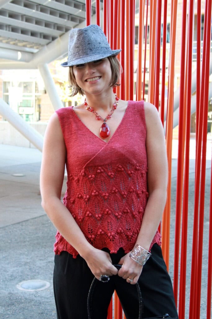 A smiling woman wearing a gray fedora, red sleeveless knit top, black pants, and bold red jewelry stands in front of vertical red poles in an urban outdoor setting. -Marly Bird