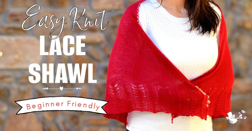A bright red knit lace shawl with an airy stitch pattern is draped over a white sweater, shown against a stone wall.