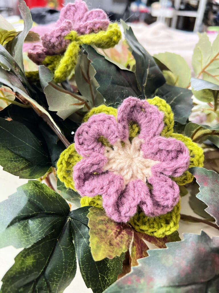 Crocheted purple and cream flowers with green leaves are nestled among realistic artificial green leaves. The background appears blurred, highlighting the detailed crochet work in the foreground. -Marly Bird