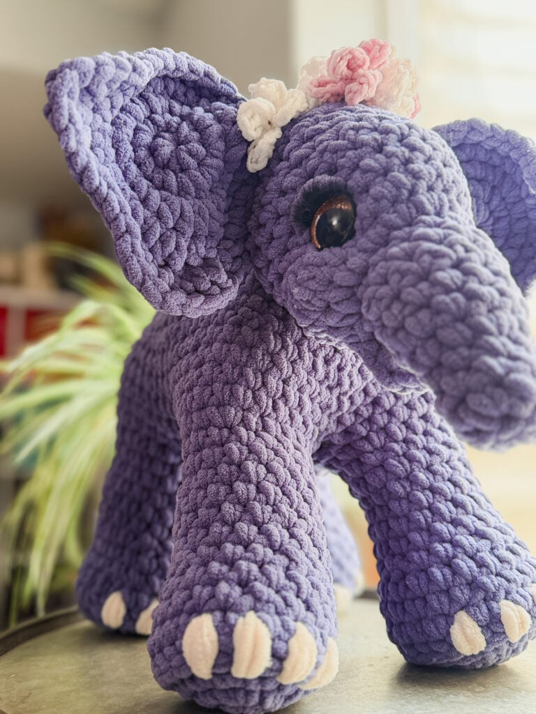 Amigurumi crochet elephant in purple yarn with big ears, white toes, crocheted floral headband, and soft textured surface.