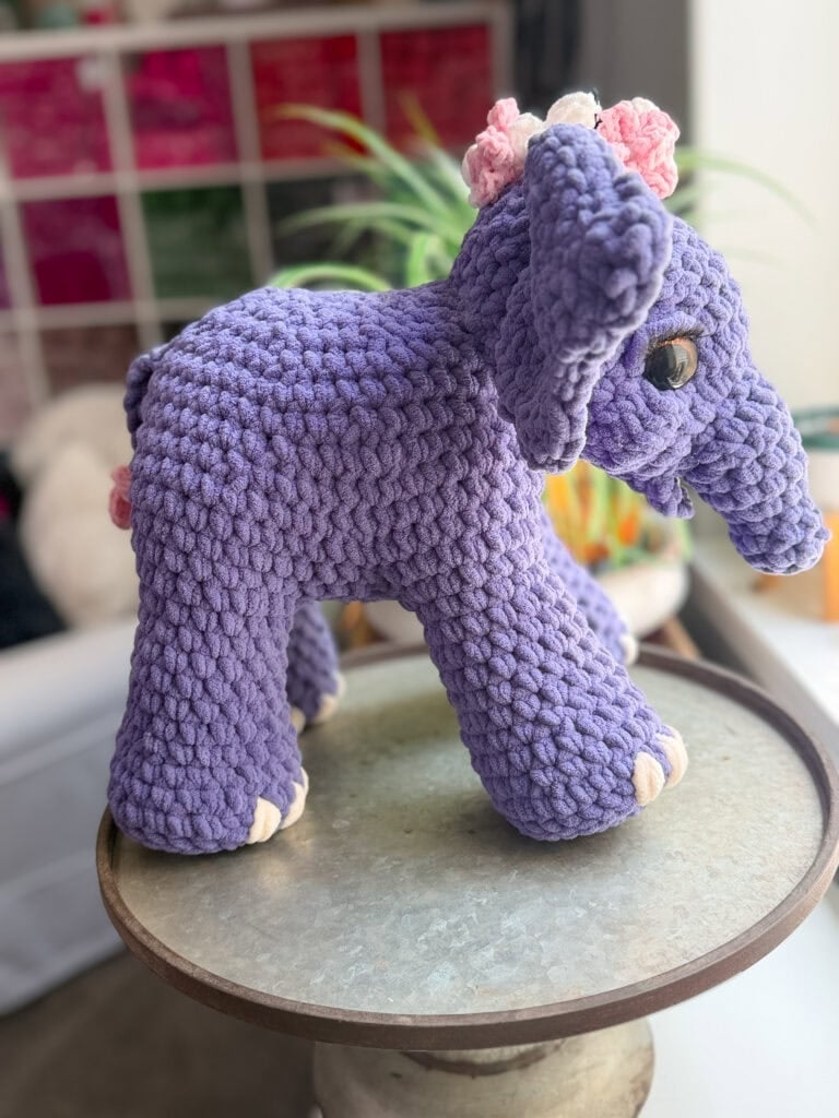 A crocheted purple amigurumi elephant with pink and white flower accents on its head sits on a table near a window.
