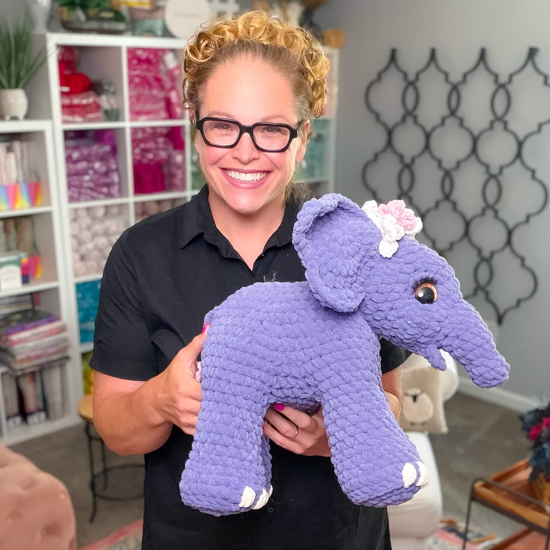 A person displays a hand-crocheted purple elephant with a pink flower, showing detailed amigurumi stitching in a yarn-filled craft room.