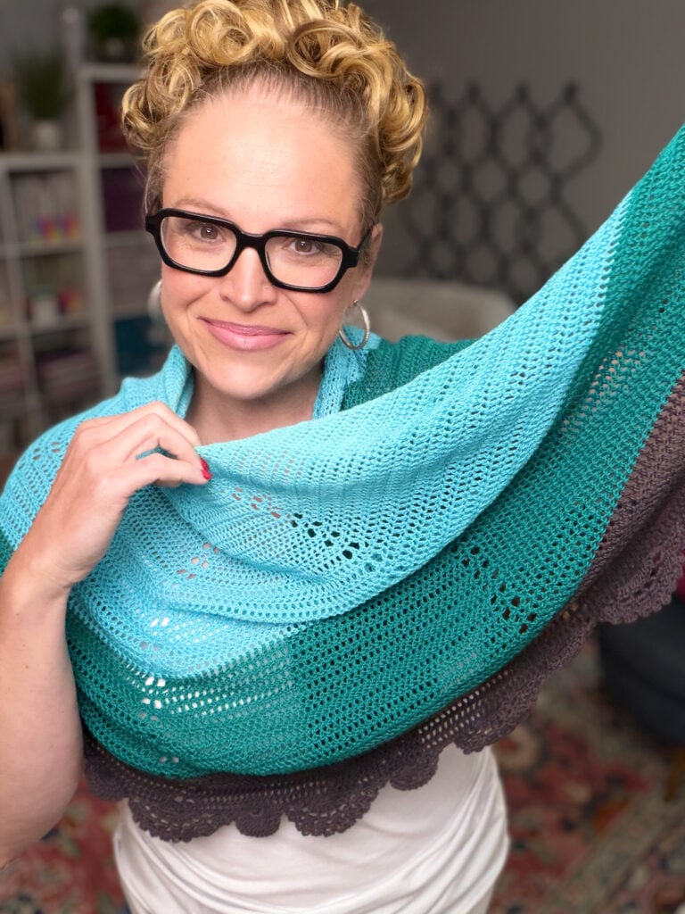Crochet Lace Shawl in blue, green, and gray displayed to show intricate stitch detail and soft texture. Shelves behind.