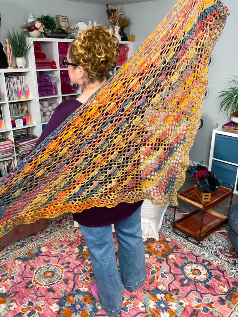 A person with curly hair, wearing glasses, jeans, and a purple top, displays a colorful Boothbay Harbor Shawl in a cozy room with shelves of yarn, books, houseplants, and a vibrant patterned rug. -Marly Bird