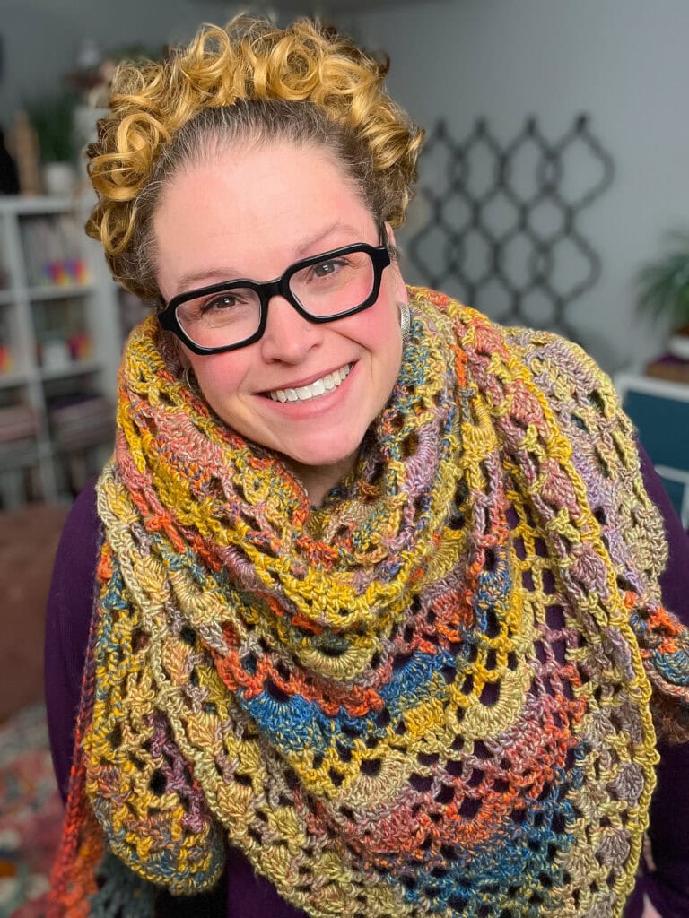 A smiling person with curly blonde hair and glasses wears a large, colorful Boothbay Harbor Shawl with intricate crochet patterns, standing indoors with shelves and decor in the background—a perfect showcase of Marly Bird’s Crochet Shawl Pattern. -Marly Bird