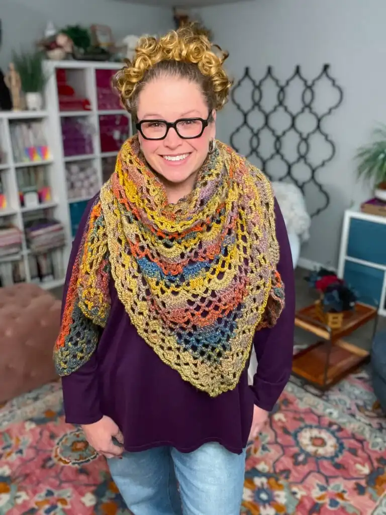 A smiling person with curly blonde hair and glasses wears a large, multicolored Boothbay Harbor Shawl over a purple top, standing in a cozy, colorful room with bookshelves and patterned rugs. -Marly Bird