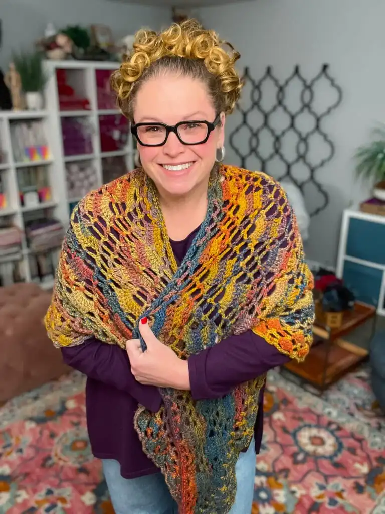 A smiling person with curly hair and glasses stands indoors, wearing a colorful Boothbay Harbor Shawl over a purple top. The background features shelves with yarn, plants, and patterned décor—perfect inspiration for your next free crochet shawl pattern. -Marly Bird