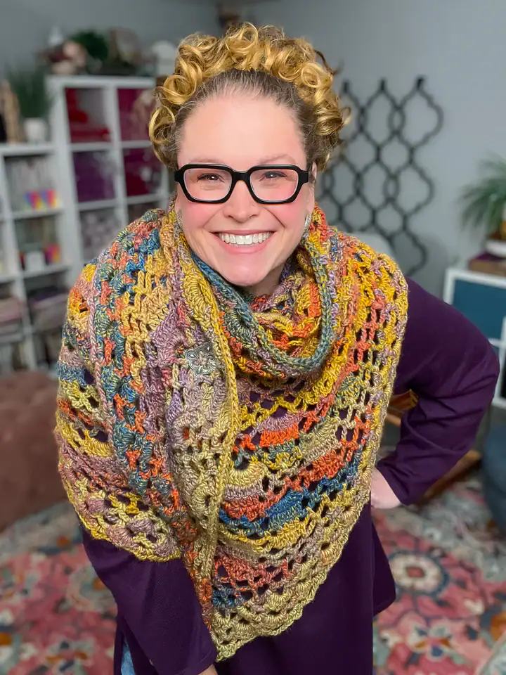 A smiling person with curly hair and glasses wears a colorful, textured crochet shawl—made from free knitting and crochet patterns—over a purple top, standing in a cozy, well-decorated room with bookshelves and plants in the background. -Marly Bird