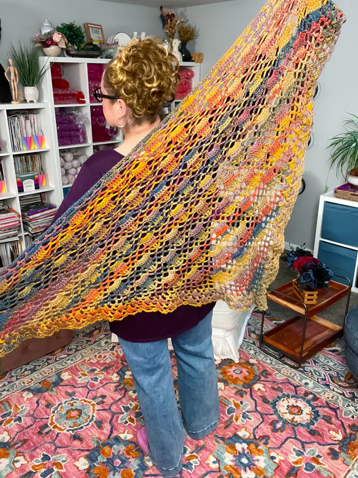 A person with curly hair in a bun models a large, colorful, hand-knit shawl spread across their back in a cozy, brightly decorated room filled with shelves, yarn, free knitting and crochet patterns, and a floral rug. -Marly Bird
