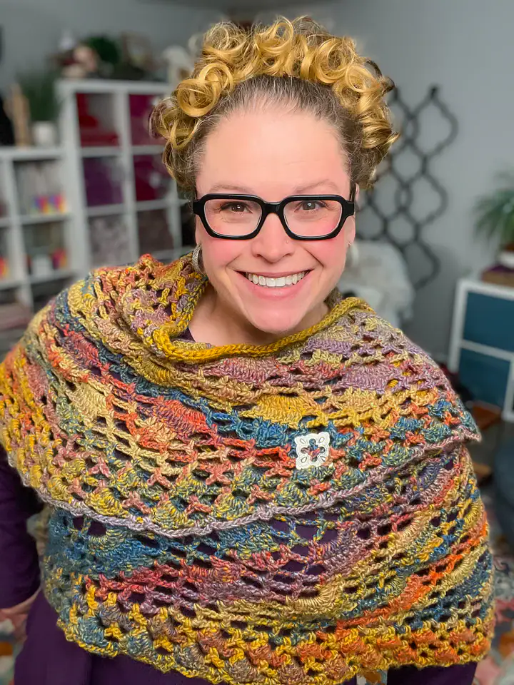 A smiling person with curly hair and glasses wears a colorful, crocheted shawl with a floral pin, standing indoors in a cozy room filled with plants and shelves—perfect inspiration for free knitting and crochet patterns. -Marly Bird