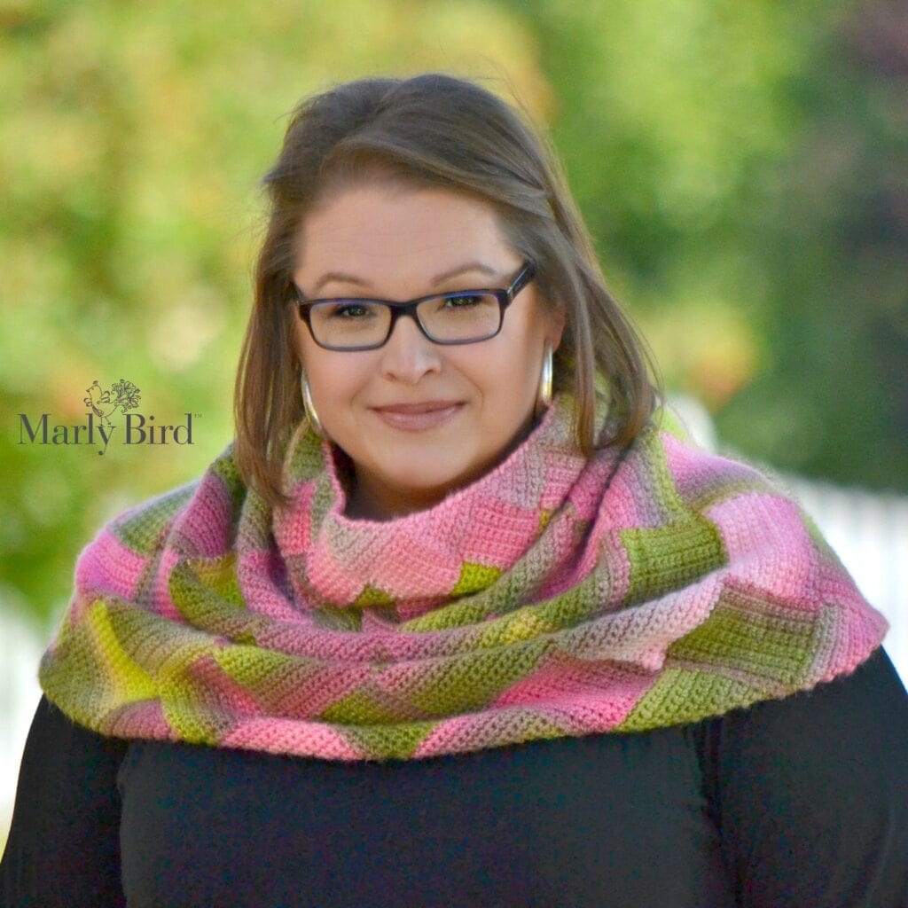 Woman models a pink and green crocheted shawl with bold text “Marly Bird” left; stitch texture visible over black top.