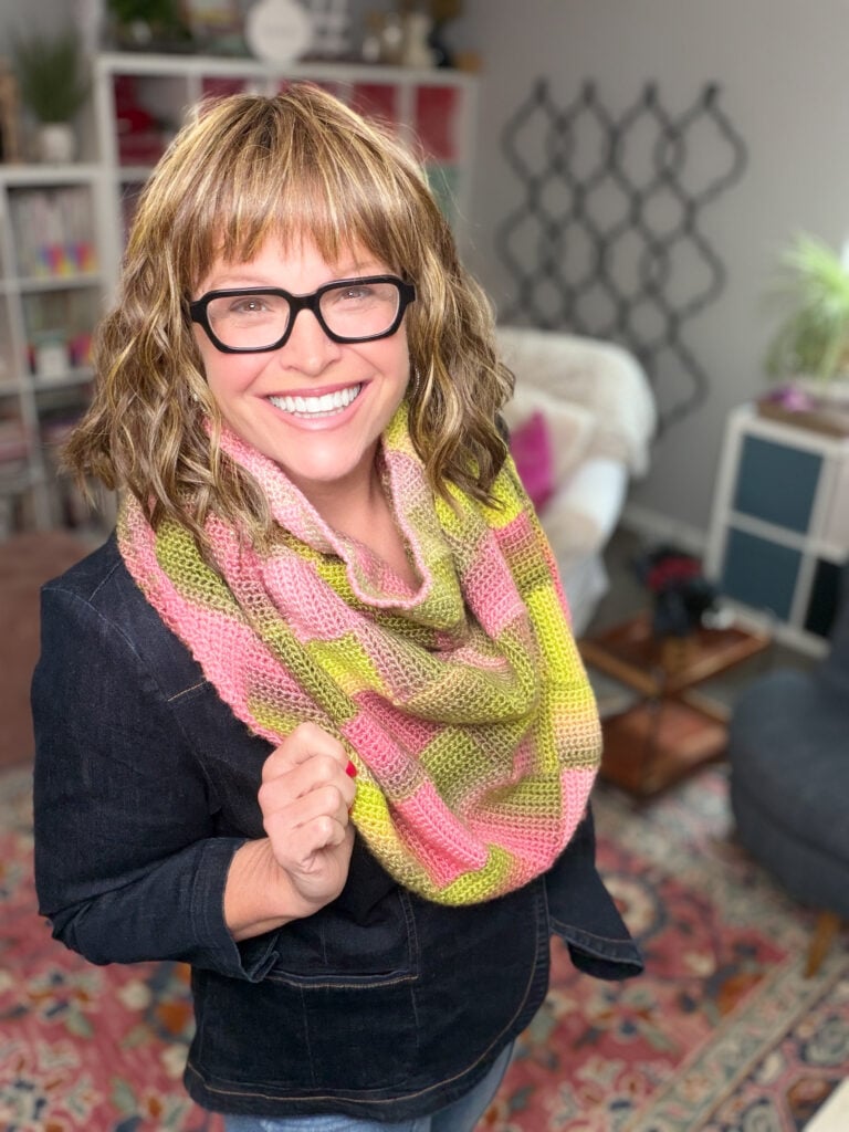Woman models a pink and yellow crochet scarf with visible textured stitches; bookshelves and home decor in the background.