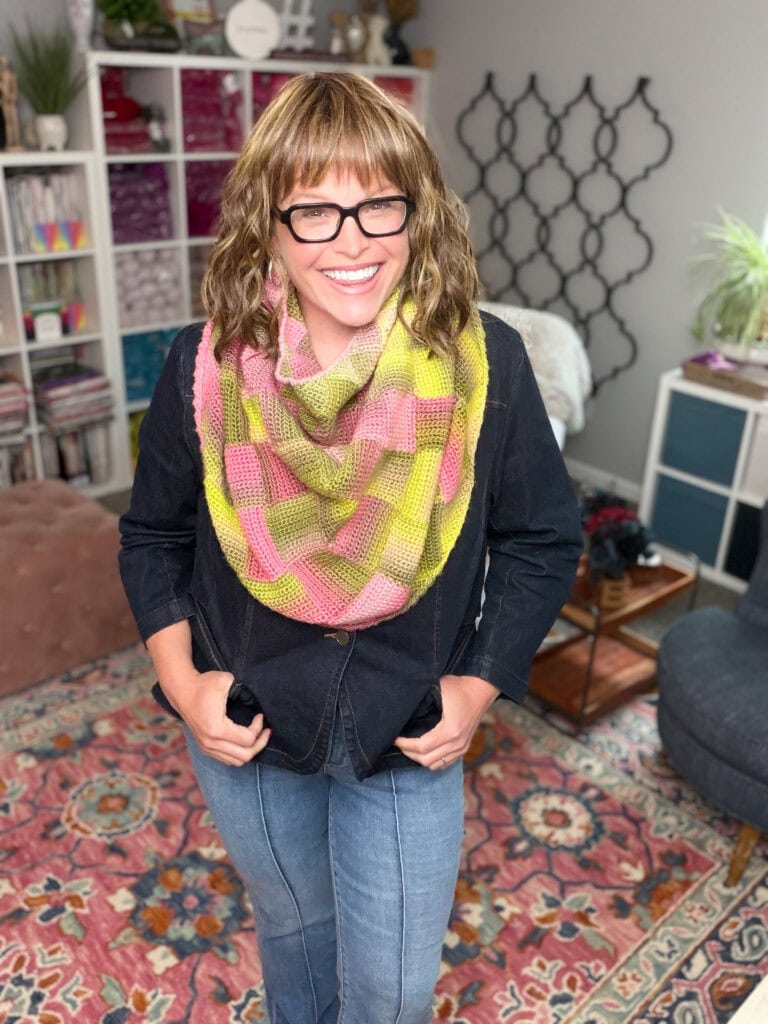 Woman models the Boysenberry Bramble Cowl, showing off its textured stitch pattern in a cozy room with bookshelves and plants.