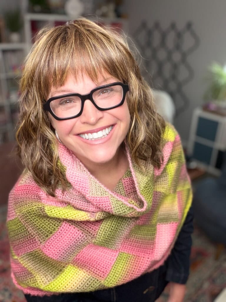 A woman wears a colorful crocheted Boysenberry Bramble Cowl, showing wavy stitch texture and drape; blurred shelves behind her.