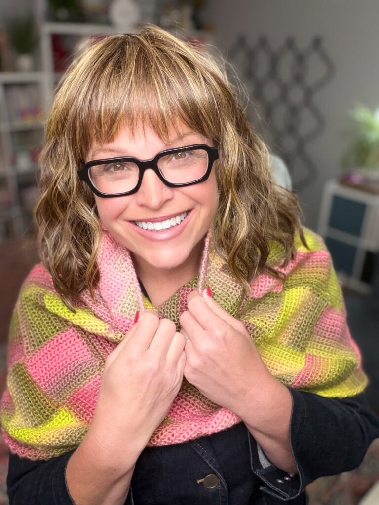 A pink and yellow Boysenberry Bramble Cowl with textured stitches, wrapped around a woman's shoulders indoors.