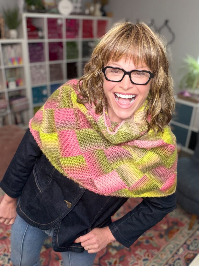 Woman wears a hand-knit, textured Boysenberry Bramble Cowl indoors, with shelves of books and yarn in the background.