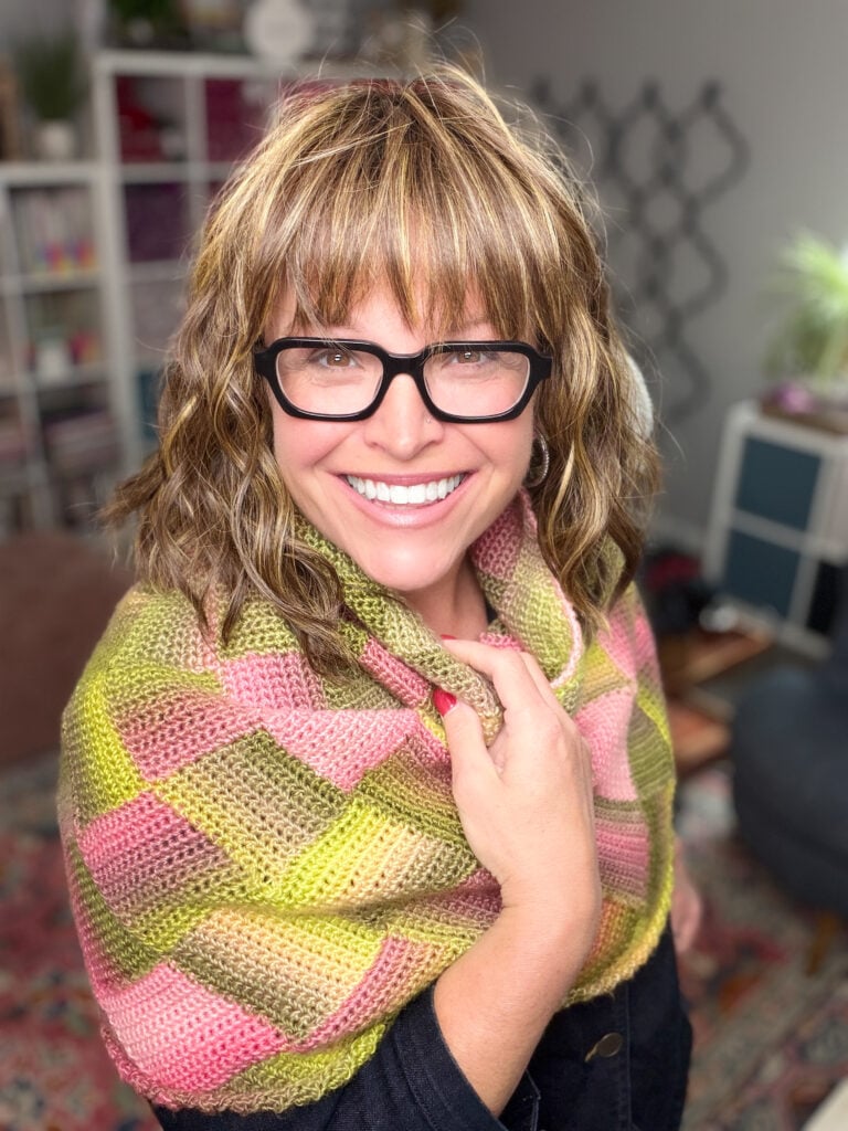 Colorful crochet cowl in greens, yellow, and pink; visible stitch detail and texture as worn by a smiling woman.