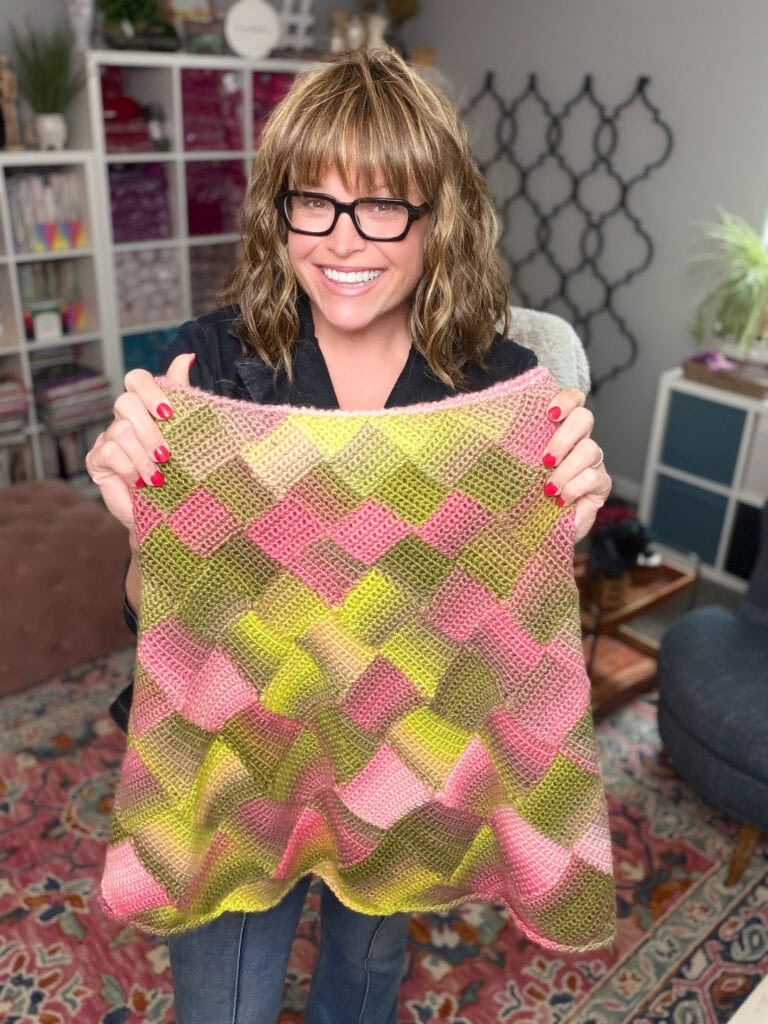 A woman displays a single crochet entrelac blanket in pink, yellow, and green checkerboard; yarn shelves fill the cozy background.
