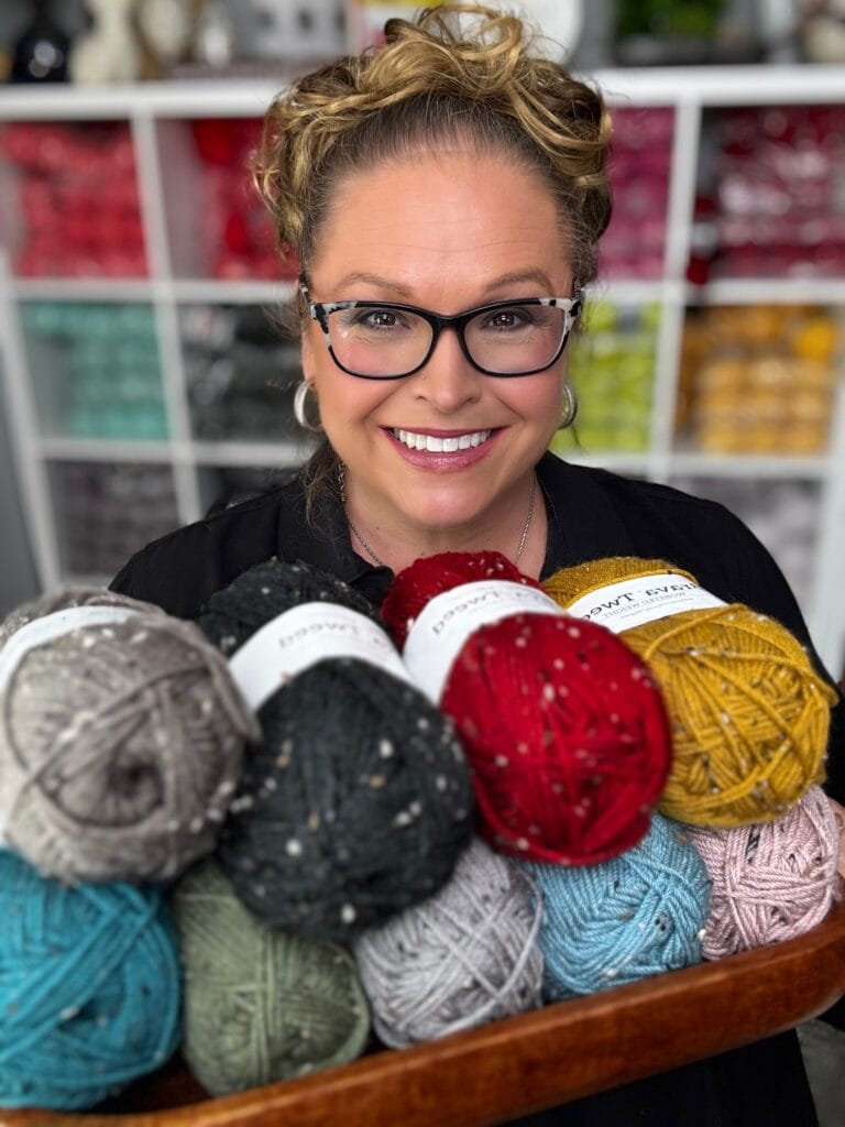 A smiling woman with glasses holds a wooden tray filled with colorful skeins of yarn, ready for the Sleigh the Holiday Make-Along 2025. Shelves with neatly arranged yarn balls are visible in the background. -Marly Bird