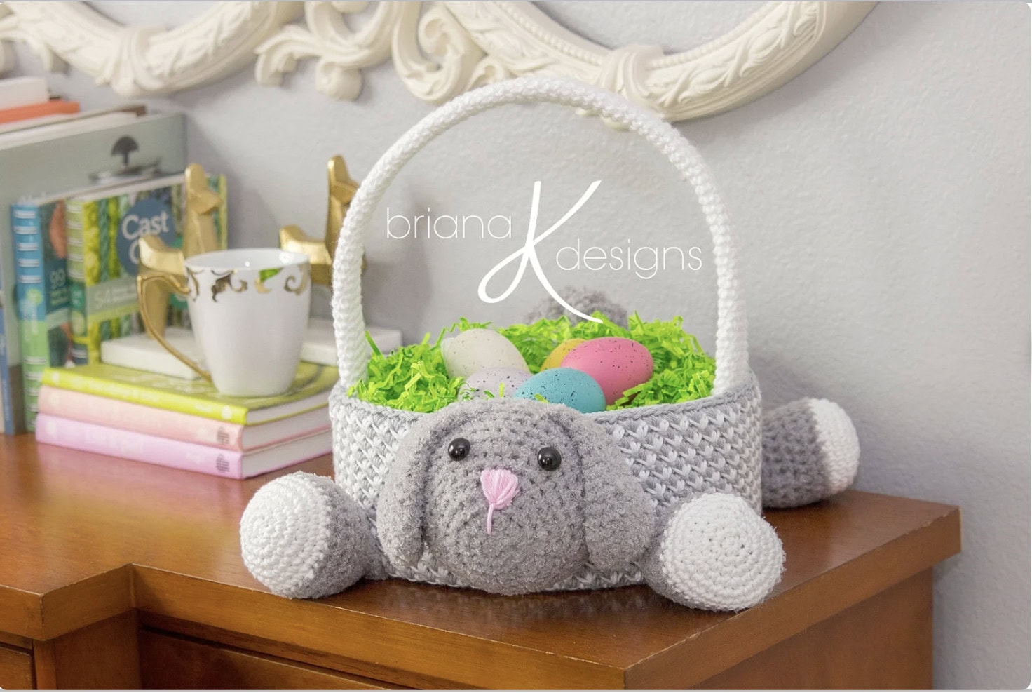 A crocheted basket shaped like a gray bunny with floppy ears and pink nose holds colorful eggs and green grass. It sits on a wooden surface in front of books and a decorative mug, beneath a white ornate mirror. "Briana K Designs" is on the image. -Marly Bird