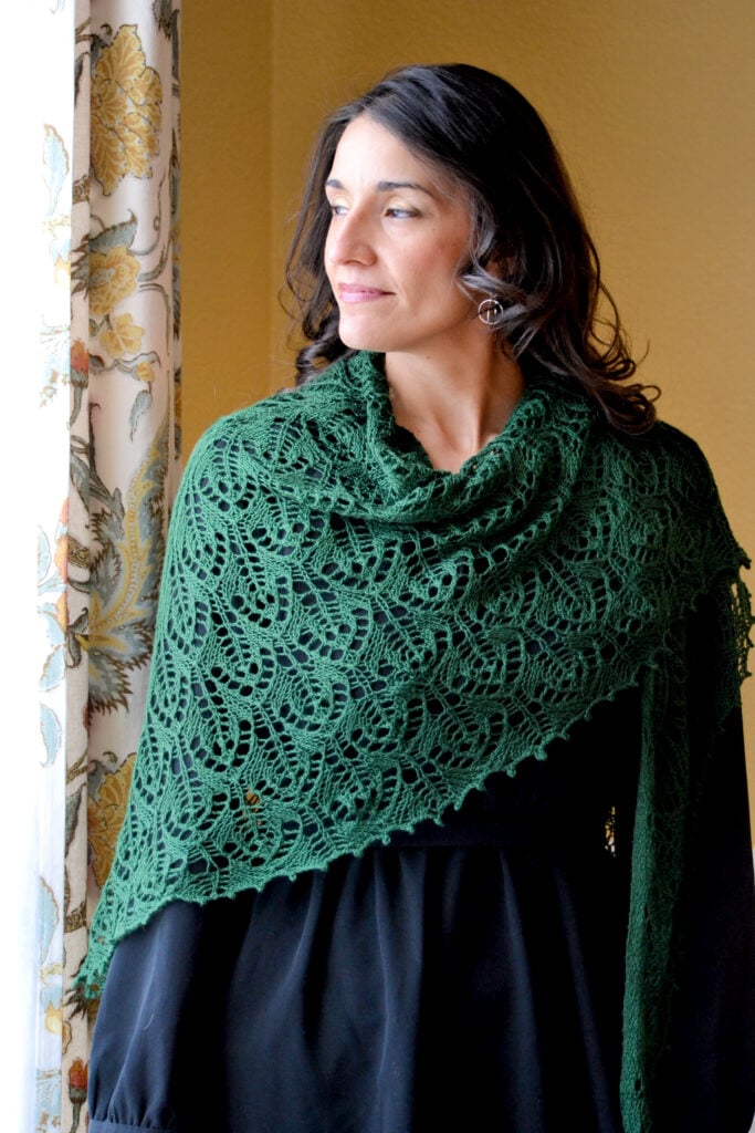 Brontë knit lace shawl in dark green jewel-tone yarn, modeled by a woman over a black dress by a window, showing delicate openwork stitch detail and drape.