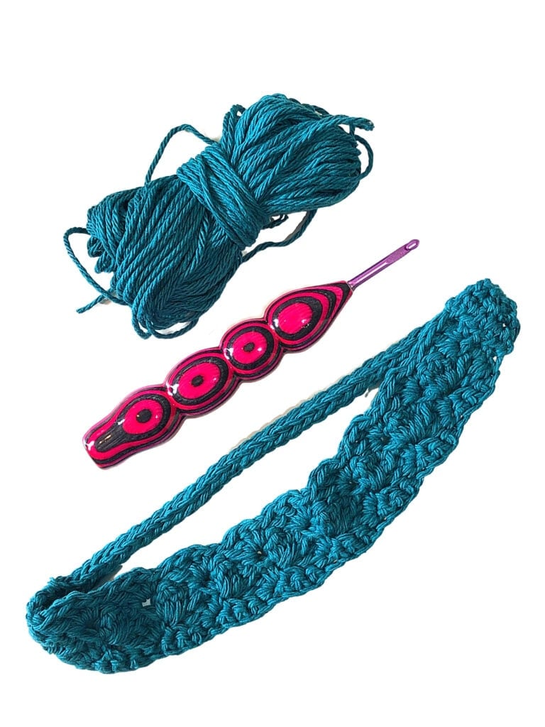 Teal yarn, a pink and black crochet hook, and a partially crocheted teal swatch on white background; visible stitch detail.